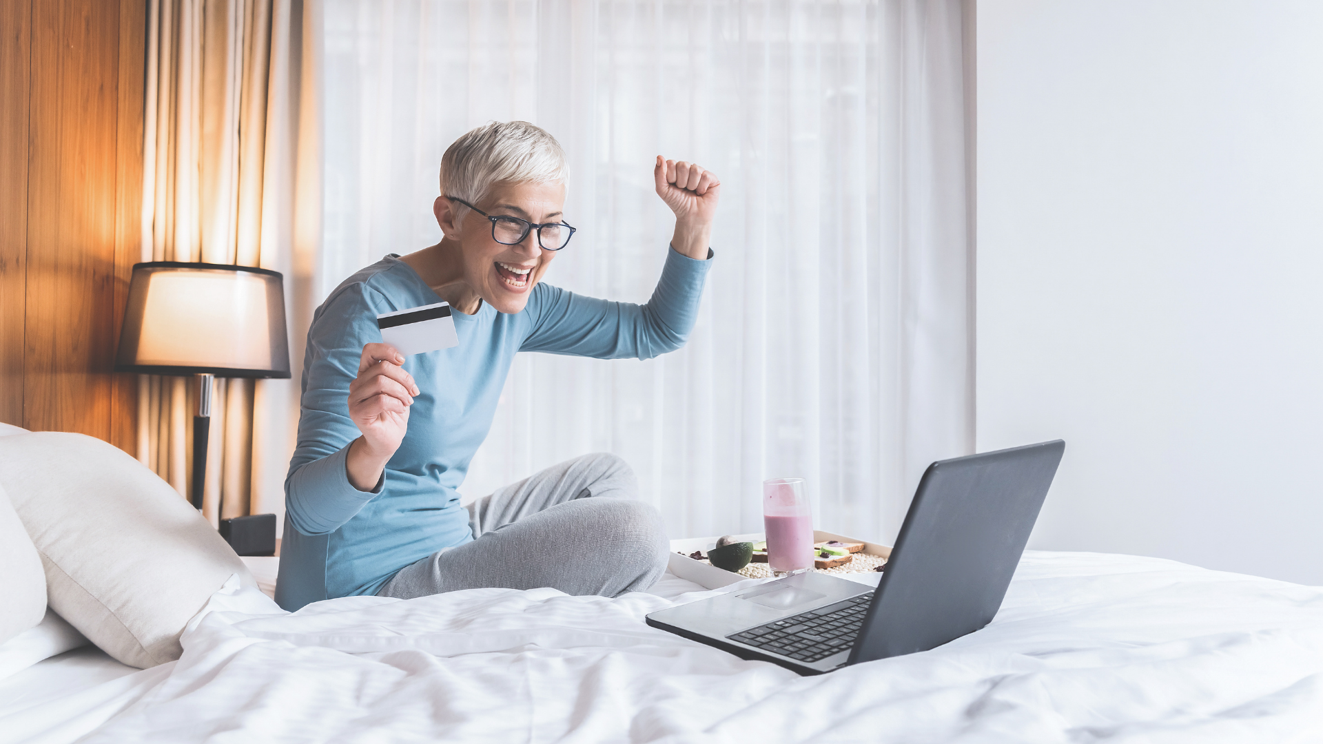 Une femme souriant sur son lit devant son ordinateur portable et tenant une carte de paiement dans la main